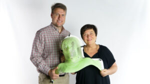 Paul and Julie are smiling, standing side by side in front of a white background. Together they are holding a large green plastic mask the size of a person's head and shoulders, called an immobilisation mask.