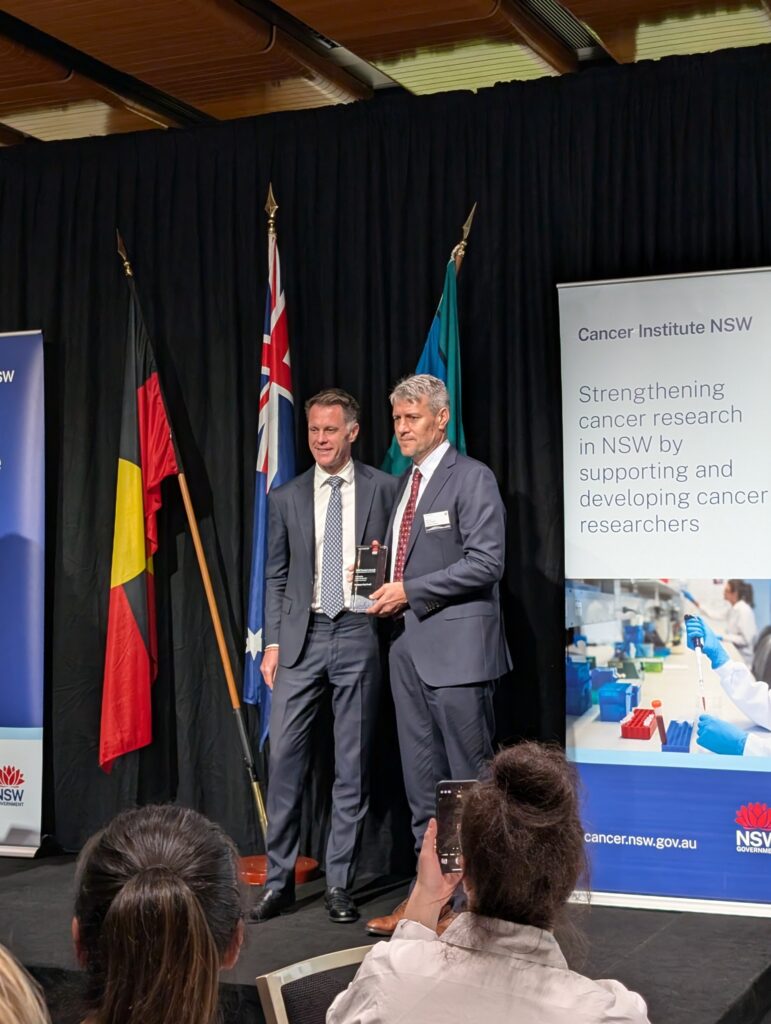 Paul Keall and NSW Premier Chris Minns are standing in front of the Australian, Aboriginal, and Torres Strait Islander flags. Paul is holding a glass award.